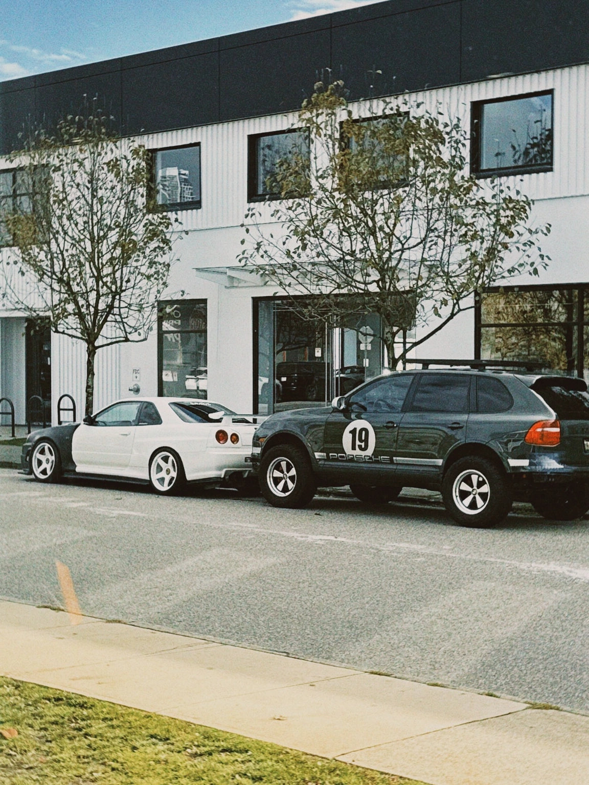 Porsche Cayenne 957 with rally number 19 decal on AkiOverland Edition W wheels, parked behind a Nissan Skyline GT-R R34