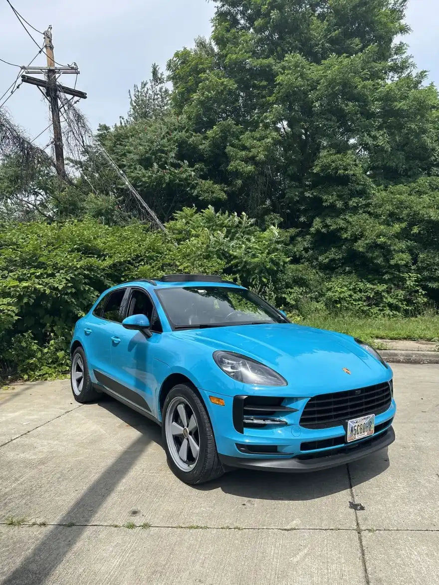 Front three-quarter view of 2021 Porsche Macan Miami Blue Sully on a Cleveland Ohio urban lot with dense summer greenery — pre-Edition W Forged Fuchs wheel build stage