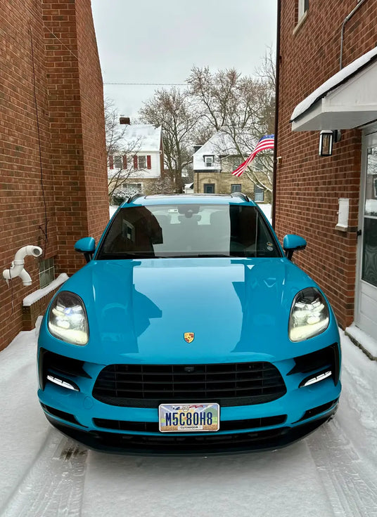 Straight front view of 2021 Porsche Macan Miami Blue Sully with headlights on in Cleveland Ohio winter snow between brick buildings — Cuyahoga County, the only colour for miles