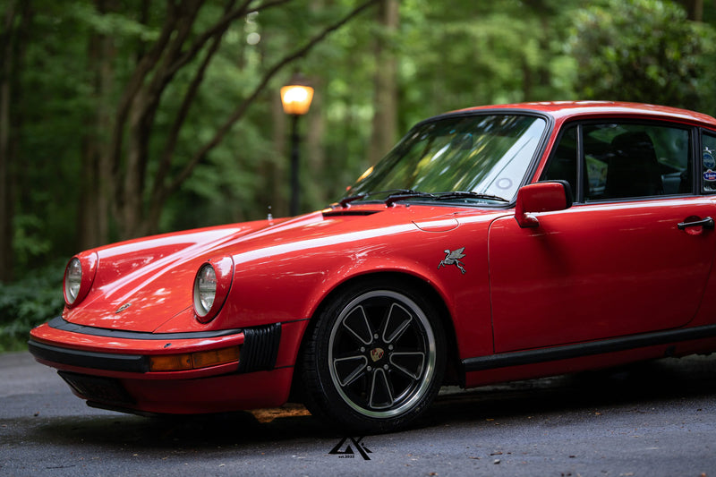 A classic red Porsche 911 coupe parked outdoors on a paved path with a blurred forest background.
