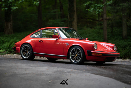 A classic red Porsche 911 coupe parked on an asphalt road with a dense green forest in the background.