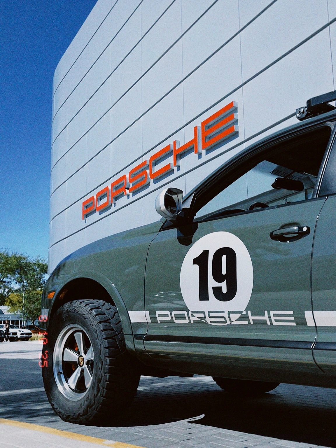 Porsche Cayenne 957 with rally number 19 decal and 5x130 wheels outside Porsche dealership.