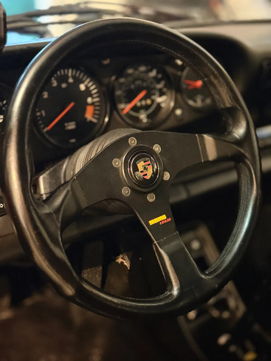 Close-up of Momo Corsa steering wheel in 1982 Porsche 911 SC with Porsche crest center cap and original tachometer visible behind
Option B