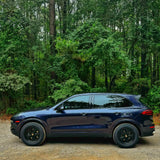 Dark metallic blue Porsche Cayenne 958 on AkiOverland Classic Fuchs wheels in satin black, parked on a gravel road in the forest
