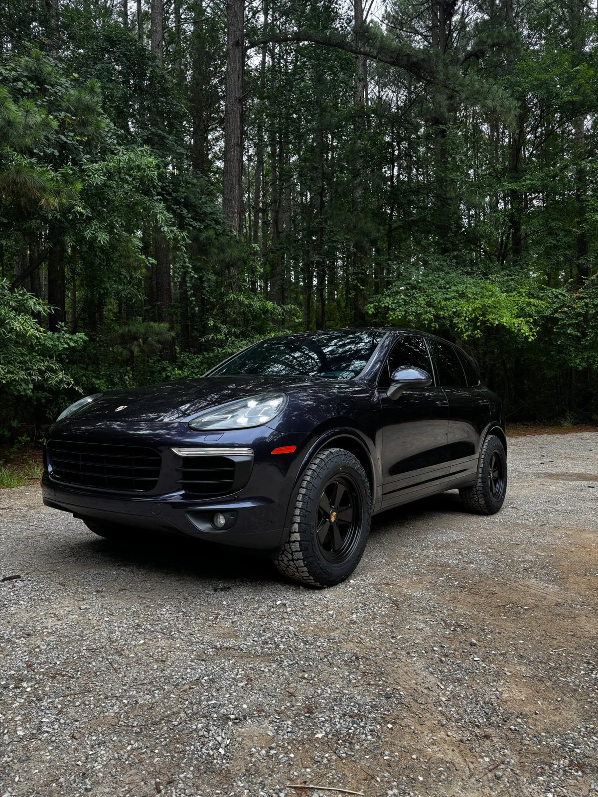 Satin black AkiOverland Classic Fuchs forged wheels on a dark blue Porsche Cayenne 958 against a forest backdrop on gravel