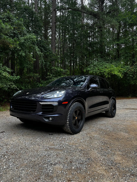 Satin black AkiOverland Classic Fuchs forged wheels on a dark blue Porsche Cayenne 958 against a forest backdrop on gravel