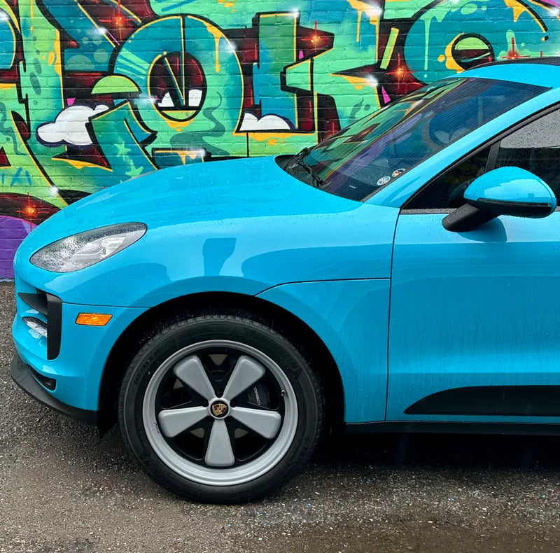 Close front quarter of 2021 Porsche Macan Miami Blue Sully showing stock Porsche wheel against Oshawa Ontario graffiti wall — before AkiOverland x WrapWorkz Edition W Forged Fuchs wheel upgrade