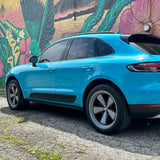 Rear three-quarter view of 2021 Porsche Macan Miami Blue Sully showing stock wheels against Cleveland Ohio tropical floral mural — before AkiOverland x WrapWorkz Edition W Forged Fuchs wheel fitment
