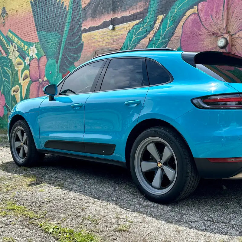 Rear three-quarter view of 2021 Porsche Macan Miami Blue Sully showing stock wheels against Cleveland Ohio tropical floral mural — before AkiOverland x WrapWorkz Edition W Forged Fuchs wheel fitment