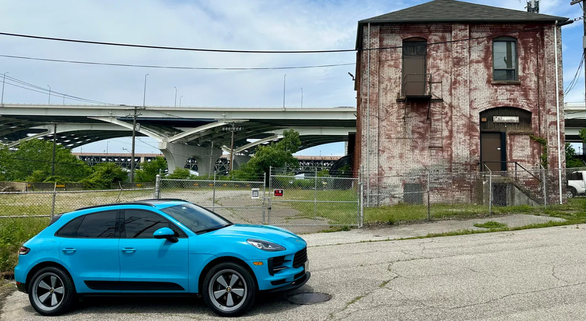 Side profile of 2021 Porsche Macan Miami Blue Sully at Cleveland Ohio industrial district near highway overpass and historic brick building — pre-AkiOverland x WrapWorkz Edition W Forged Fuchs wheel build