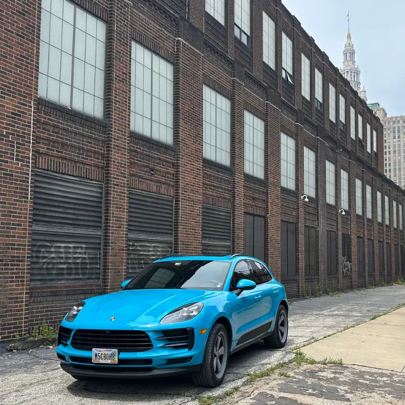 Front view of 2021 Porsche Macan Miami Blue Sully parked against historic Cleveland Ohio brick warehouse with Terminal Tower visible in background — pre-Edition W Forged Fuchs build