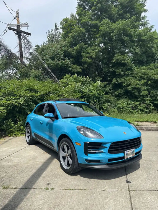 Front three-quarter view of 2021 Porsche Macan Miami Blue Sully on a Cleveland Ohio urban lot with dense summer greenery — pre-Edition W Forged Fuchs wheel build stage